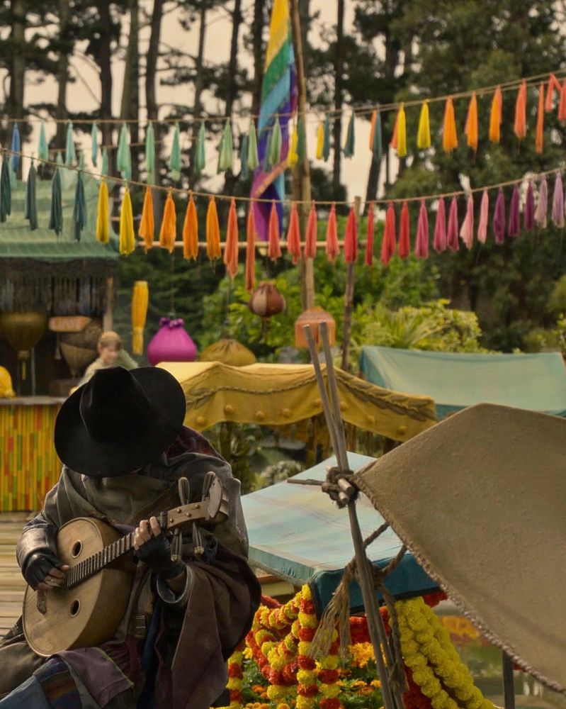 Still of Ned Low in disguise sitting with his head down at the floating market in season 2 episode 6 of Our Flag Means Death. He’s wearing a black hat, a dark poncho, and playing a stringed instrument. Behind him there’s a rainbow flag, as well as hanging colourful tassels.