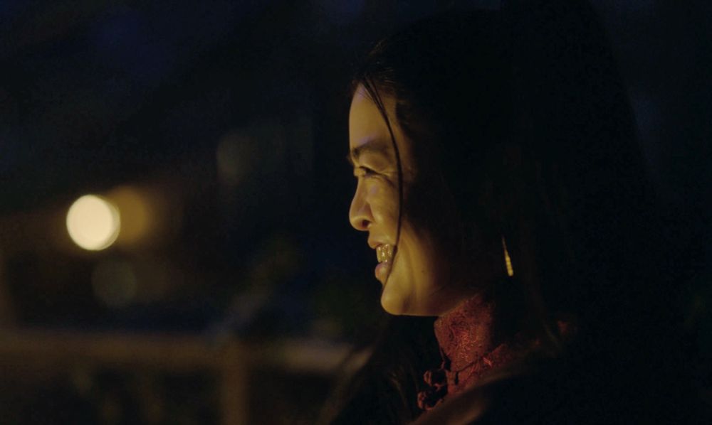 Closeup of Zheng sitting in profile across the table from Oluwande (not in frame) in season 2 episode 7 of Our Flag Means Death. It’s night and there’s a warm glow on her face from a nearby candle. She’s looking over at Olu with heart eyes and the biggest smile.