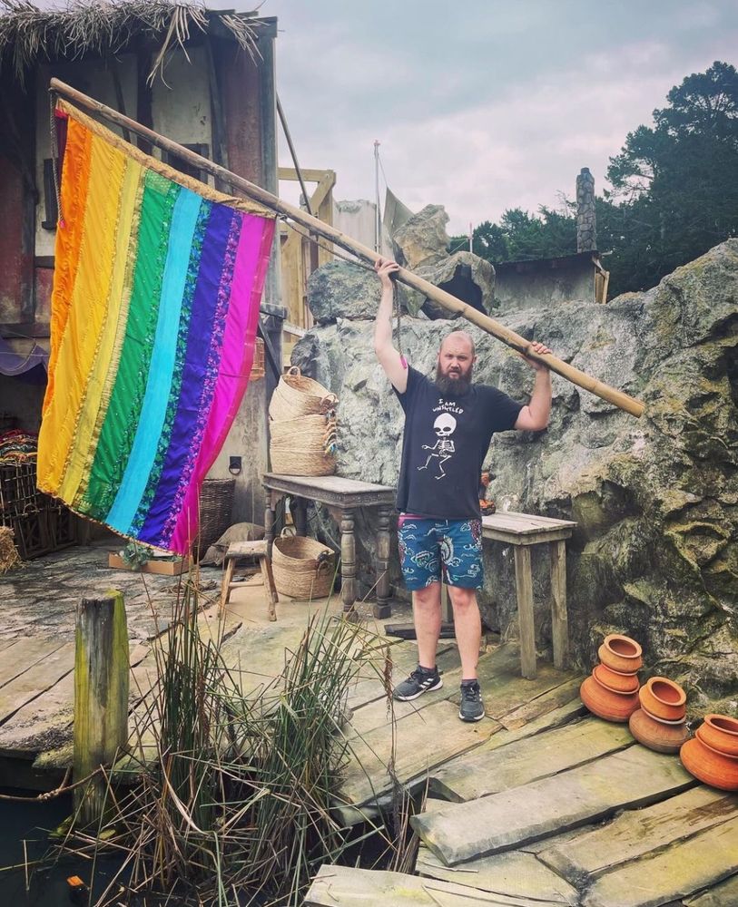 BTS shot of the rainbow flag from season 2 episode 6 of Our Flag Means Death. It’s on a wooden pole and being held up at an angle by a guy in a t-shirt and shorts. Image is by set decorator, Megan Vertelle (megsetdec on insta).