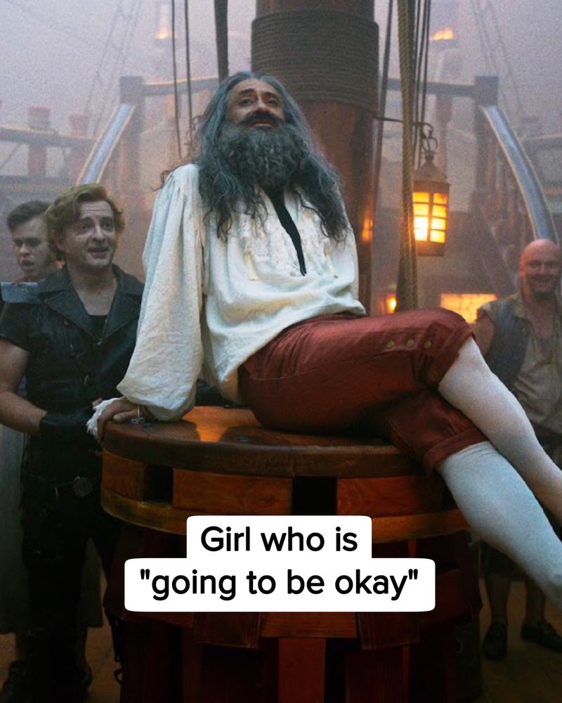 Close shot of Ed Teach in Stede’s clothes sitting on the capstan with his legs crossed (Season 1 episode 4 of Our Flag Means Death). He’s leaning back a bit and looking up with a confident expression. The crew stand behind him. Near the bottom of the image is black text against a white background, it says: Girl who is “going to be okay.”
