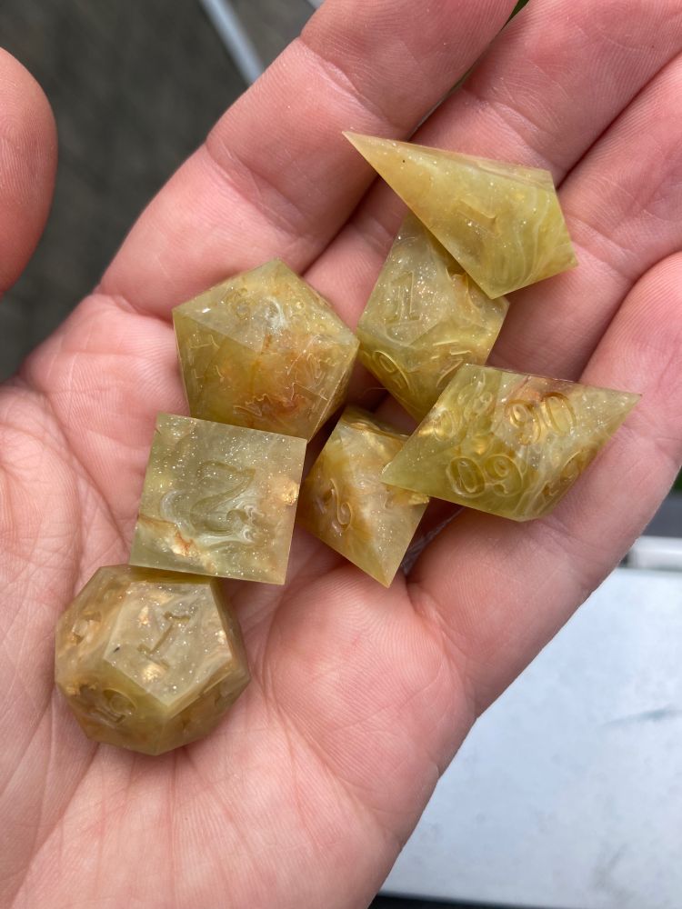 An open palm holds a matte, unfinished set of polyhedral dice. Brown and white wisps swirl through a base of spring yellow-green, gold leaf and glitter peeking through even in this unpolished state.