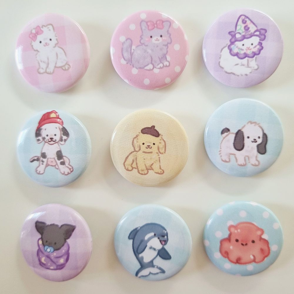 A photo of a grid of 3 by 3 mini badges. On the top row are all cat badges: a white kitten on a pink gingham background, a purple kitten on a pink polkadot background, a cat clown cat on a purple background. On the middle row are three puppy badges: a dalmatian pup in a firefighter hat on a blue background, a yellow puppy on a yellow background, a white puppy on a blue background. At the bottom is a bat badge on a purple background, a baby orca on a blue background and an orange dumbo octopus on a blue background