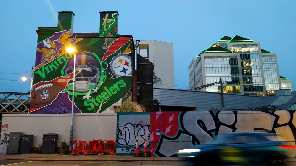 Landscape photo at dusk of the view across Dublin's Tara Street to a wall and above it to two upper stories of the side of a pub where there used to be a giant 3-dimensional mural/sculpture, made of found metal and other items, of a squirrel which now has a 2-dimensional mural celebrating the upcoming NFL game in Dublin between the Vikings and the Steelers. The wall is plain crams with traffic boxes, orange barriers and then completely covered in graffiti on the right where above it behind the pub is the long grey structure that is Tara Street Railway Station and behind it a clump of pyramid topped small glass skyscrapers (at a stretch).