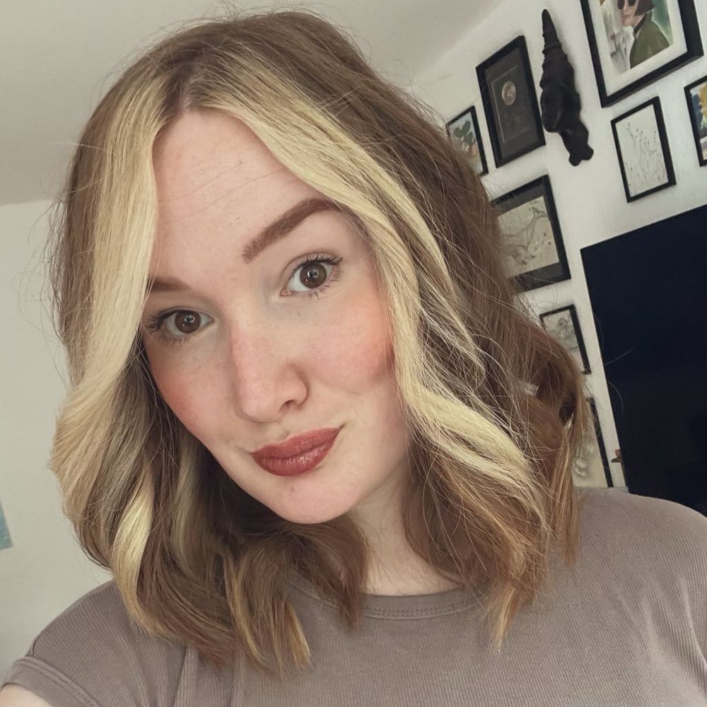 Man sieht ein Selfie von mir. Ich hab kastanienfarbende gelockte Haare mit vorne zwei sehr hellblonde Strähnen drin. Im Hintergrund ist noch ein wenig meine Bilderwand zu sehen.