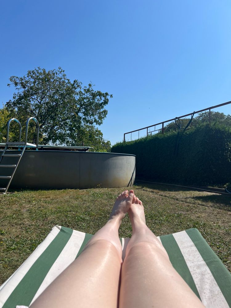 Beine auf einer gestreiften Liege, die auf einem leicht verdorrten Rasen steht. Im Hintergrund steht ein Pool. 