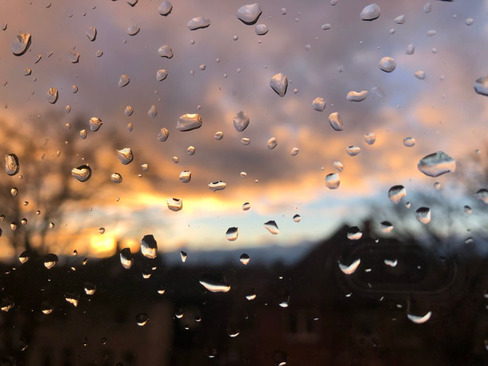 Fokussiert sind im Vordergrund die Regentropfen an der Scheibe. Im Hintergrund sieht man verschwommen einen Sonnenuntergang mit schön gefärbten Wolken. Man sieht außerdem verschwommene Häuser und Bäume.