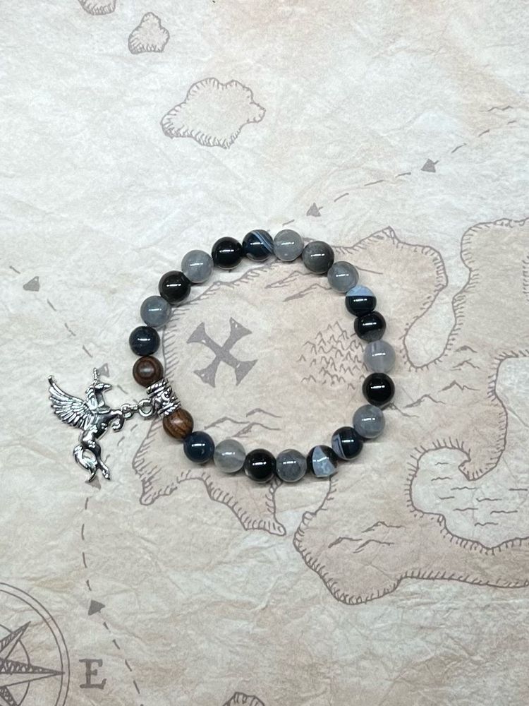 A bracelet inspired by the character Izzy Hands from Our Flag Means Death. The bracelet is made of black and grey beads. It also has two wooden beads flanking a unicorn charm.