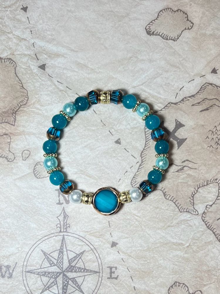 A beaded bracelet made with teal beads and pearls and gold accents inspired by the character Stede Bonnet from Our Flag Means Death.
