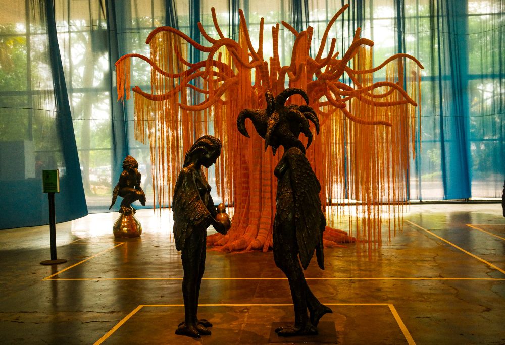 mesma obra que a anterior, porém de outro ângulo. duas figuras, a de uma garota-passaro de cabelos trançados e asas baixas segurando o mesmo "ovo" que o personagem anterior, de cabeça baixa, frente a uma mulher-passaro mais velha, com asas grandes e os cabelos formando "spikes" na cabeça dela chamando muita atenção.