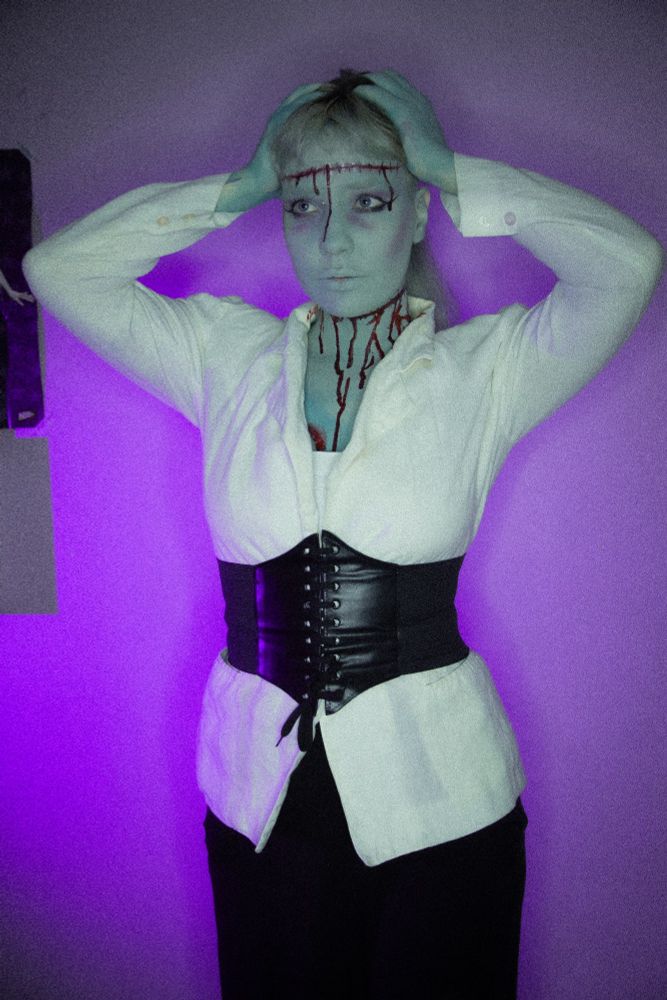 Eu, pintada de verde, com um blazer branco, corset preto e calça preta, maquiagem simulando corte na testa e no pescoço e sangue artificial escorrendo. Fundo roxo. Estou com as mãos segurando a cabeça, como se sentisse a costura da minha testa.