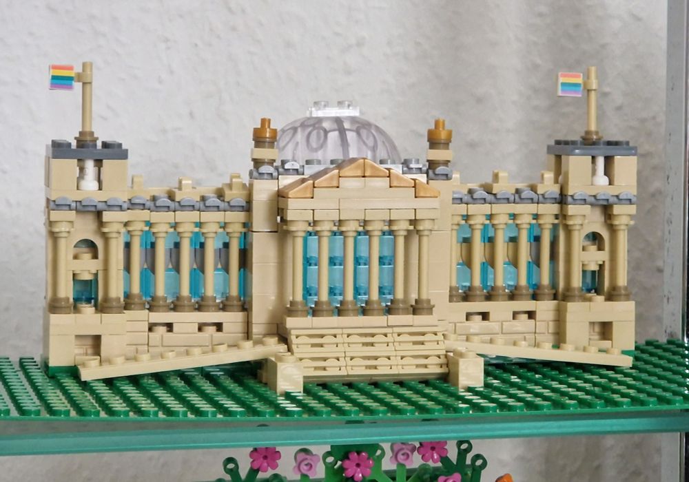 Ein Reichstagsgebäude aus Klemmbausteinen. Das Gebäude ist beige mit beige-farbenen Säulen und hellblauen Fenstern, der Rasen ist grün. Auf den Türmchen des Gebäudes rechts und links sind 2 Regenbogenfahnen.