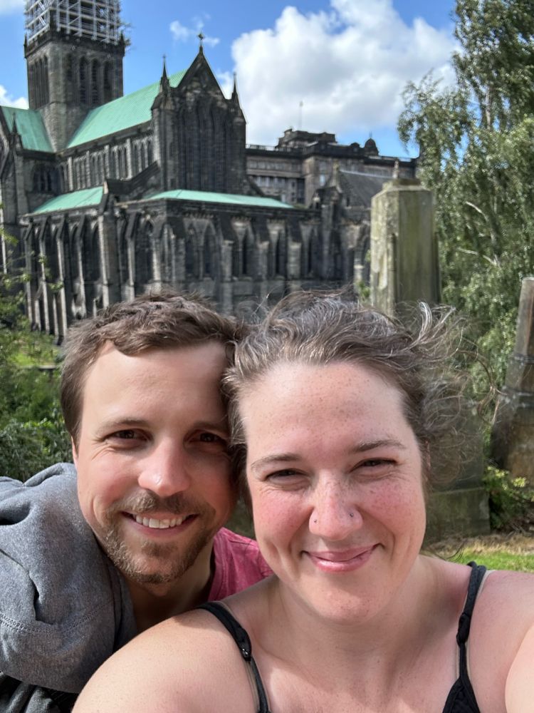Bree and partner take a selfie from the top of the path of the necropolis.  Behind them are grave markers framing the overlook of the Glasgow Cathedral. Both people are smiling.