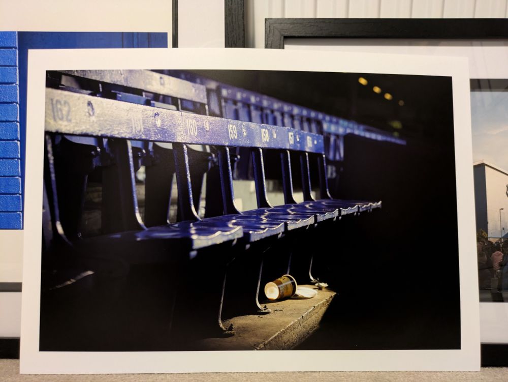 In the foreground is a print of a Pete Regan photograph of wooden Goodison seats. Behind to the left is a Gary Lambert piece. Behind to the right is a Lewis Guy piece.