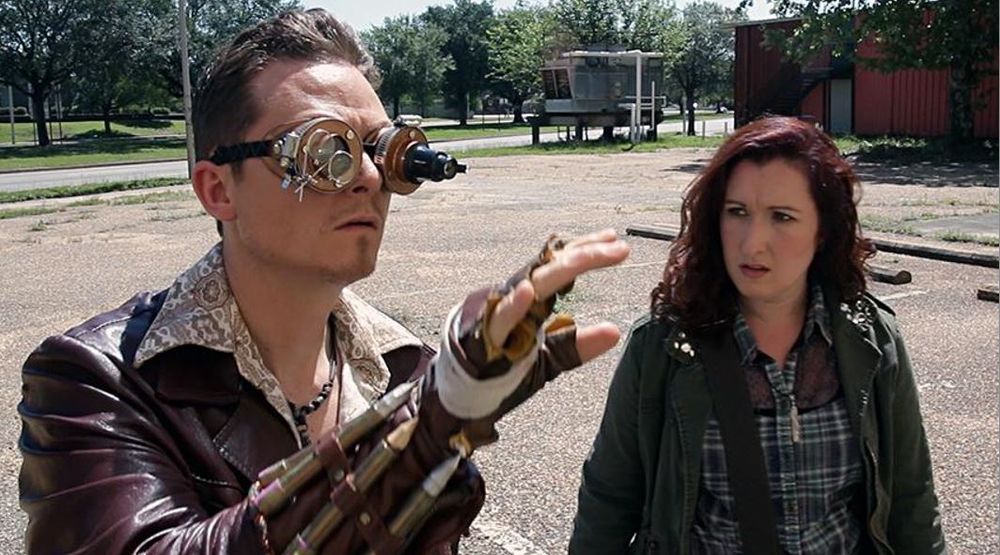 A still from Demon Squad. Khristian Fulmer is wearing a leather jacket, steampunk goggles and some kind of weapon strapped to his forearm. Erin Lilley is wearing ordinary street clothes and looking at him with concern.