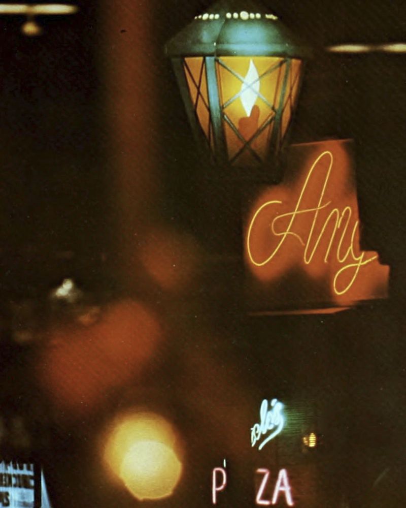 Photo taken outside with a lit streetlamp in foreground. Neon signs and lights scatter, including a bright red sign that says "pizza." From 1967 Athena yearbook.
