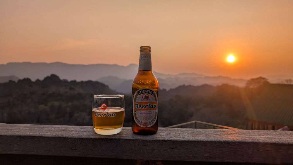 Eine Flasche und ein Glas Beerlao stehen auf einer Holzbrüstung im Vordergrund. Im Hintergrund erstreckt sich eine malerische Berglandschaft unter einem orangefarbenen Sonnenuntergang, der den Himmel und die Umgebung in warmes Licht taucht.