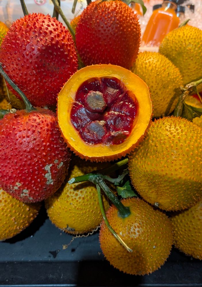A pile of spiky gac fruits in shades of orange and red, with one fruit cut open to reveal its dark red pulp and seeds, displayed on a market stand.