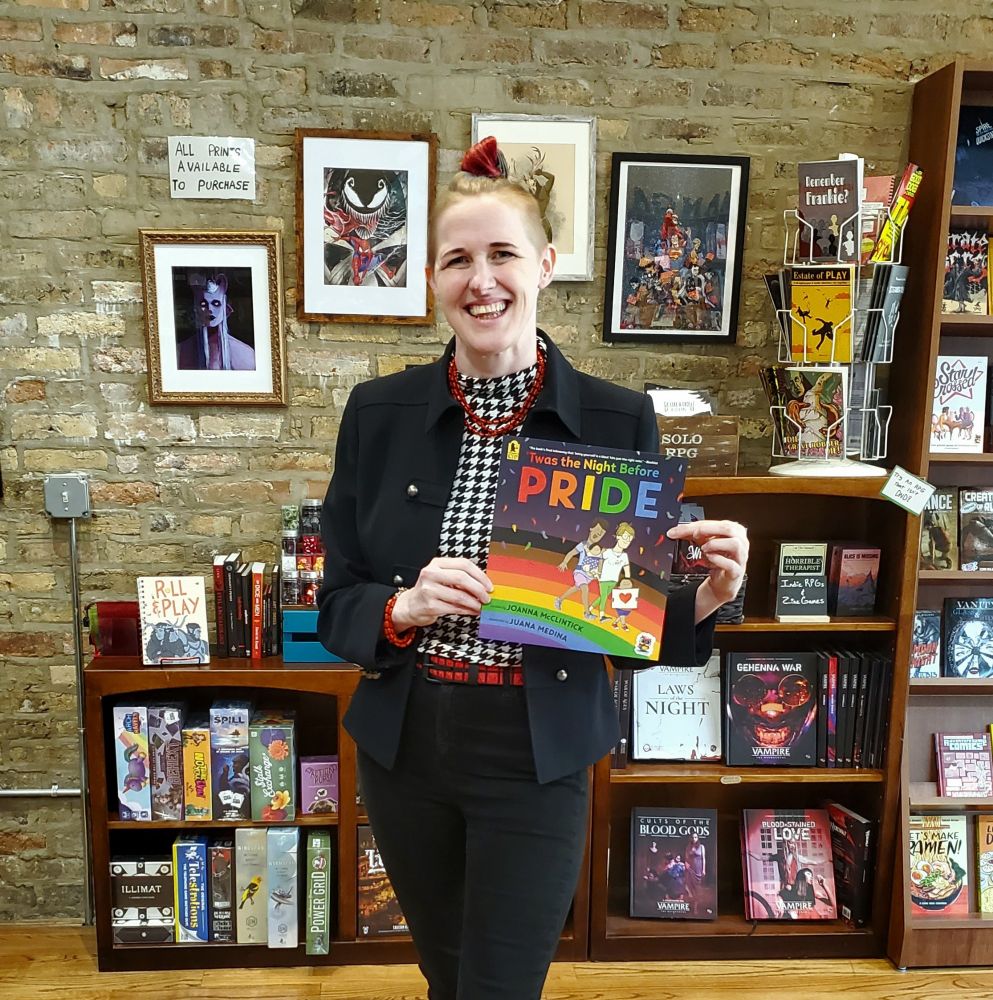 Ryvre holding a book titled the Night Before Pride in front of the game shelves at Goblin Market 