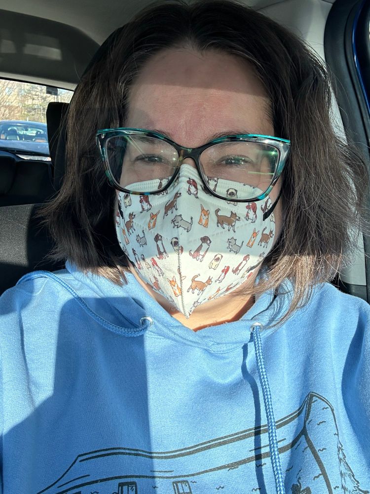 A goofy white lady in a white mask and covered in cartoon dogs looking at the camera. She is wearing a blue sweatshirt and blue glasses and is being blinded by the sun.