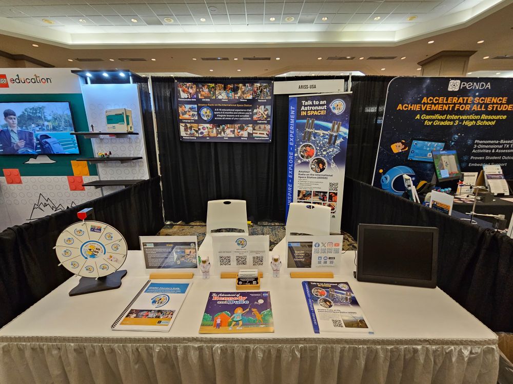 The booth prominently features ARISS-USA (Amateur Radio on the International Space Station), showcasing its program that enables students to communicate directly with astronauts aboard the ISS via amateur radio. Central displays include a detailed poster highlighting the ARISS initiative, with images of students engaging in live Q&A sessions and descriptions of 4–6-month lesson plans integrating language arts, math, science, and physical education. On the table, an ARISS Educator’s Guide, Talk to an Astronaut in Space! pamphlets, and QR codes provide resources for educators to implement the program. Small astronaut figurines and a spinning wheel with space-themed icons encourage interactive engagement, emphasizing ARISS’s mission to inspire and educate through real-world space communication.

