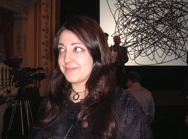 A woman wears a black embroidery jacket and has long dark hair and eyeliner