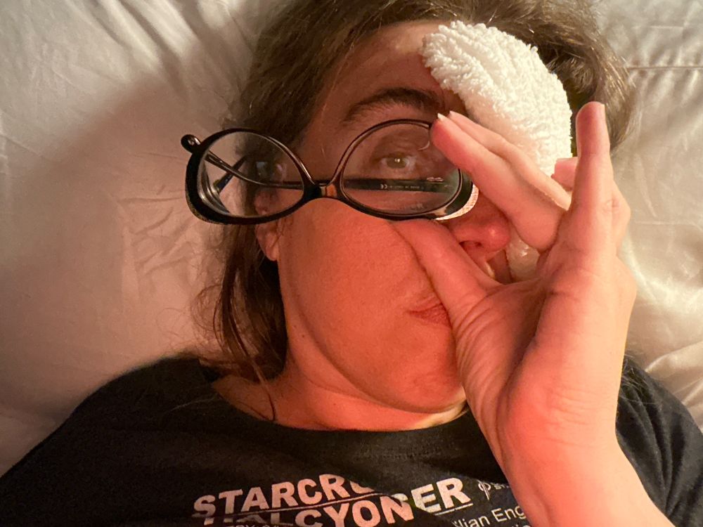A woman has a white washcloth over one of her eyes and holds it in place with her hand and in her fingers she’s holding a pair of eyeglasses upside down so that she can see out of one side.