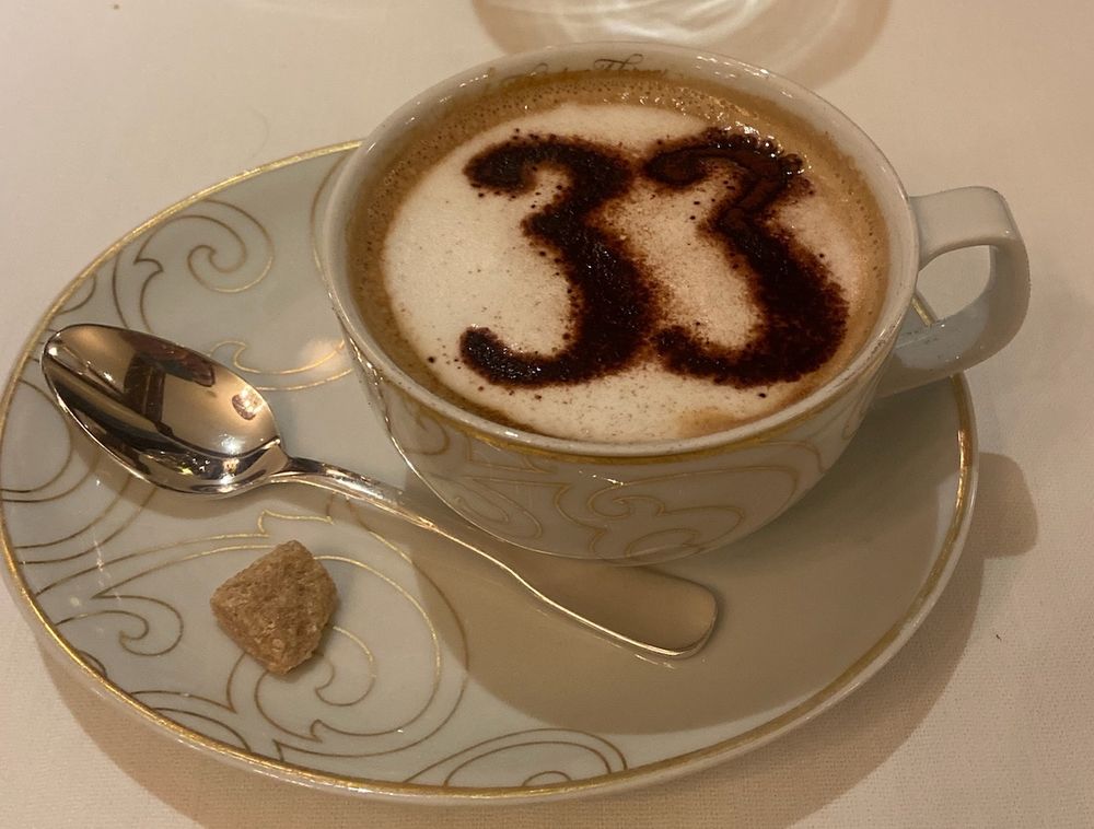 A cappuccino with cocoa on top with the numbers “33.” Served in a delicate cup with a saucer, a spoon, and a single sugar cube. 