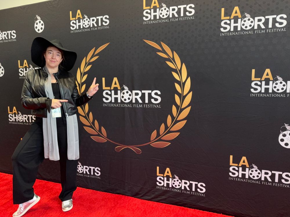 A woman in a black hat, black clothes, and silver shoes gestures toward a LA Shorts logo