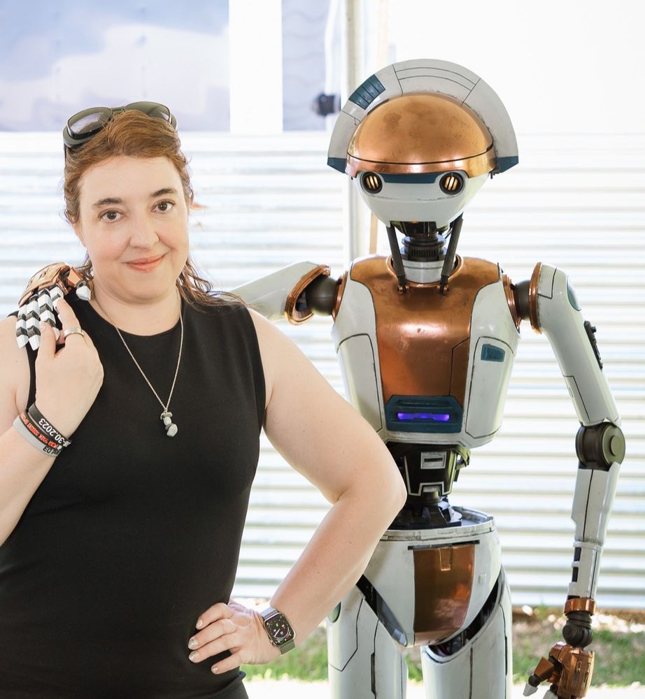 A woman has a droid’s hand wrapped around her- the droid is d3-09 from the Starcruiser halcyon