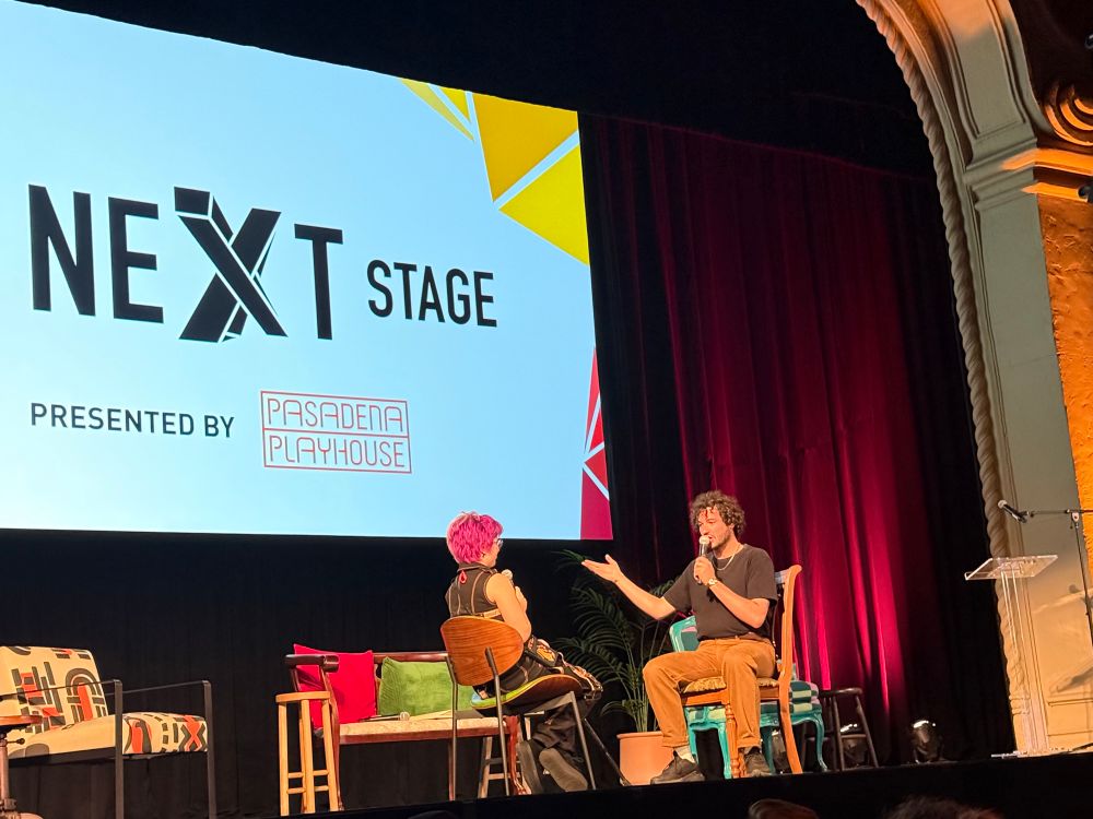 Jack Aldisert, a white brunette man gestures while speaking to a pink haired person, Cass Bailey, which enacting a small section of his work The Manikins in front of a stage audience. 