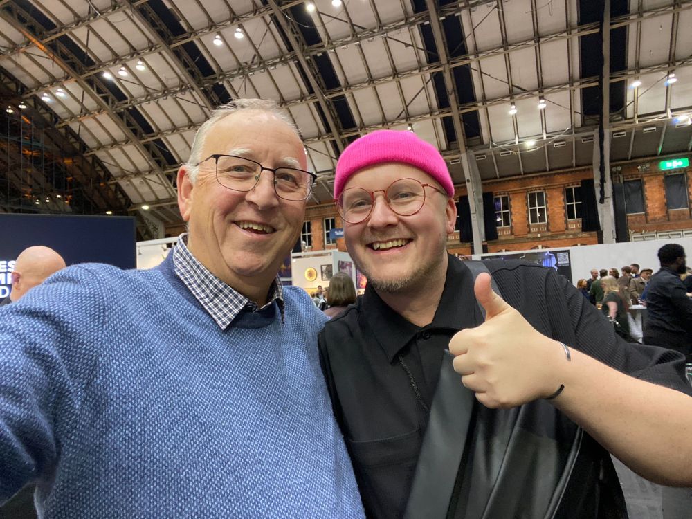Two Caucasian men stood next to each other hugging.
On the left is photographer Stuart Linden Rhodes wearing a light blue jumper and check shirt with white hair and thin black glasses. On the right is artist Garth Gratrix wearing a black plisse shirt with black leather bumbag across their chest, they also are wearing thin glasses but in vivid red and a neon pink beanie hat. They are giving a thumbs up to the camera.