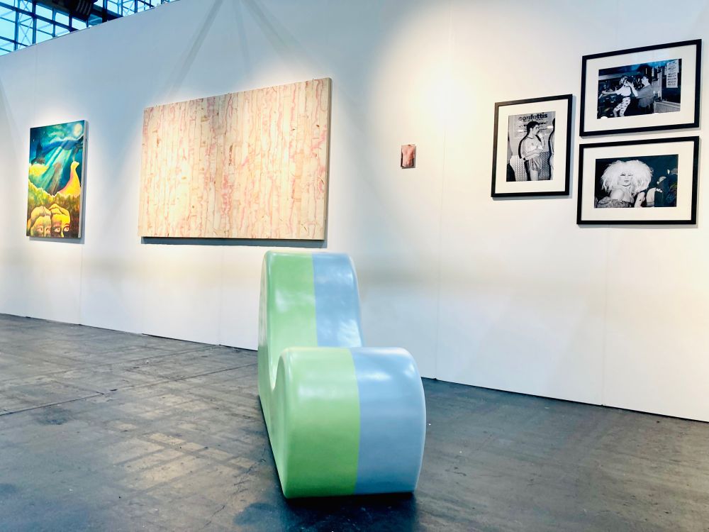 The image shows a collection of works installed in an Art Fair booth. The booth is white and floor is black.
In the foreground and centred in the image is a curved chaise like form on the floor. A sculpture by Garth Gratrix. To the left are two other artists work. A painting by Abi Hampsey showing Forest of Bowland landscape, two female face with eyes open and looking to the right at a Grouse in the landscape. The colours are bold and rich with a beam of painted light across the canvas from right to left. 
The painting next to that is by Tom Ireland and shows a number of wooden slats assembled together in a larger rectangle on the wall, each with different layers of the plywood surface sanded down to reveal various colours and textures (predominantly raw wood and red in tones). The the right of the central floor sculpture is then a small postcard size photograph by Kris Canavan depicting a torsos with the Word BumBoy scarred into the skin. Then next to this are three black frames containing photographs by Stuart Linden Rhodes. Each depict scenes archiving 90’s Queer nightlife in Manchester and Blackpool.