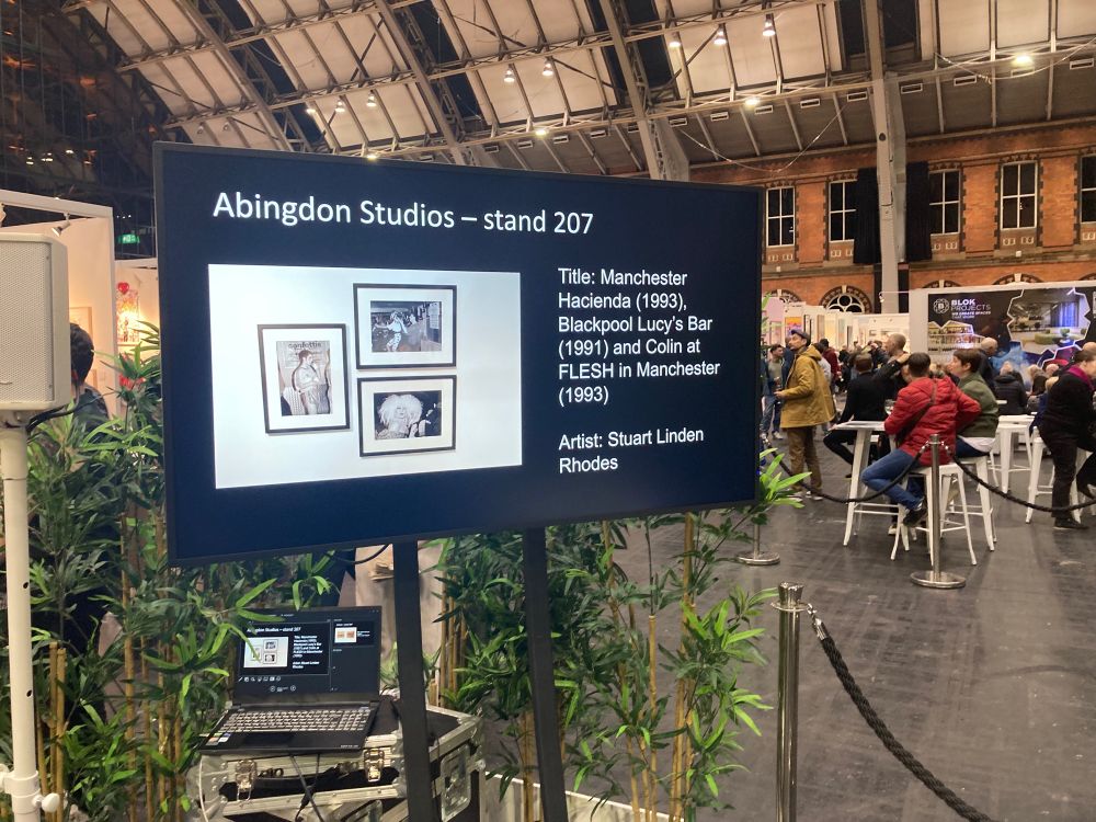Image depicts a large TV monitor installed in a bit conference scale space. The screen shows a picture of Stuart Linden Rhodes three framed photographs depicting 90’s Queer nightlife. Alongside text that reads 

Abingdon Studios STAND 228
Title: Manchester Havienda (1993)
Blackpool Lucy’s Bar (1991) and Colin at FLESH in Manchester (1993).

Promoting to an audience that these artworks have been collected by Manchester Art Gallery