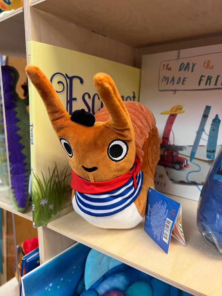 Photo of a brown plushie snail in a striped shirt, red neckerchief, and beret on a bookshelf