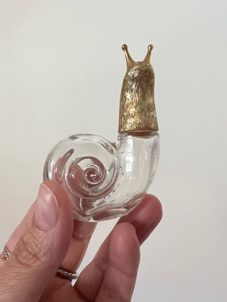 A photo of a hand holding a small empty glass perfume bottle in the shape of a snail with a metallic gold head