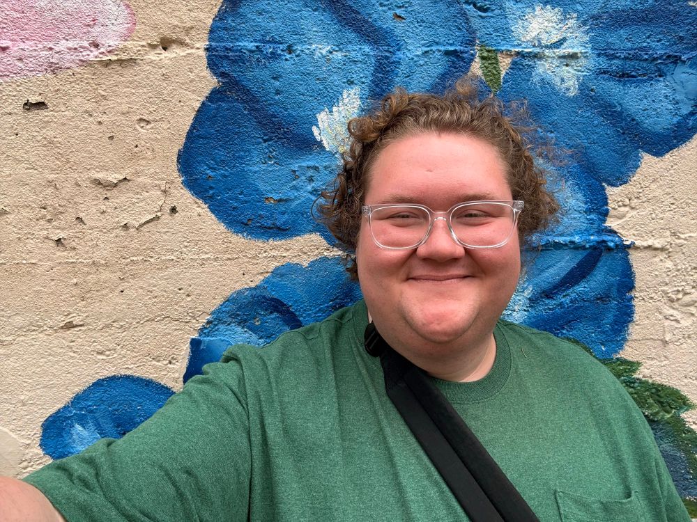 Image of me in a green shirt, smiling in front of a concrete wall adorned with painted flowers.