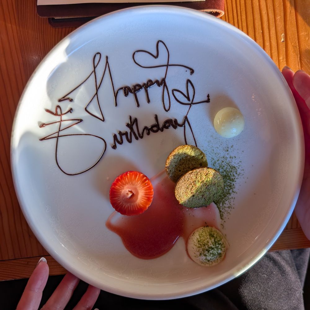 Photo of a plate with a strawberry, a coulis, a truffle and cake with 'happy birthday' written in chocolate 