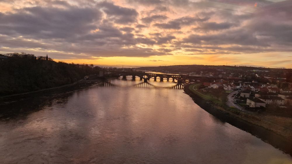 a distance view of a sunrise behind a bridge over a river, the sky is starting to turn orange and yellow