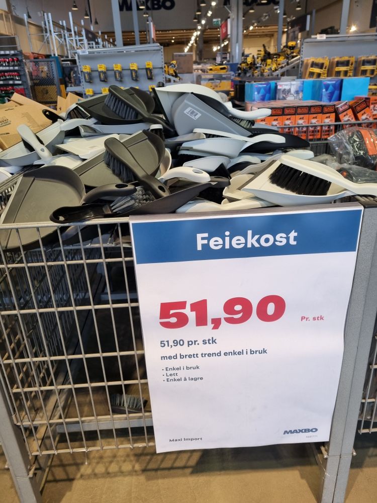 Et digert varehus i bakgrunnen. I forgrunnen står en stor metallkurv med feiekoster og tilhørende feiebrett. På kurven er det festet en plakat hvor det står
"Feiekost, 51,90 pr stk
Med brett trend enkel i bruk
-Enkel i bruk
-Lett
-Enkel å lagre" 