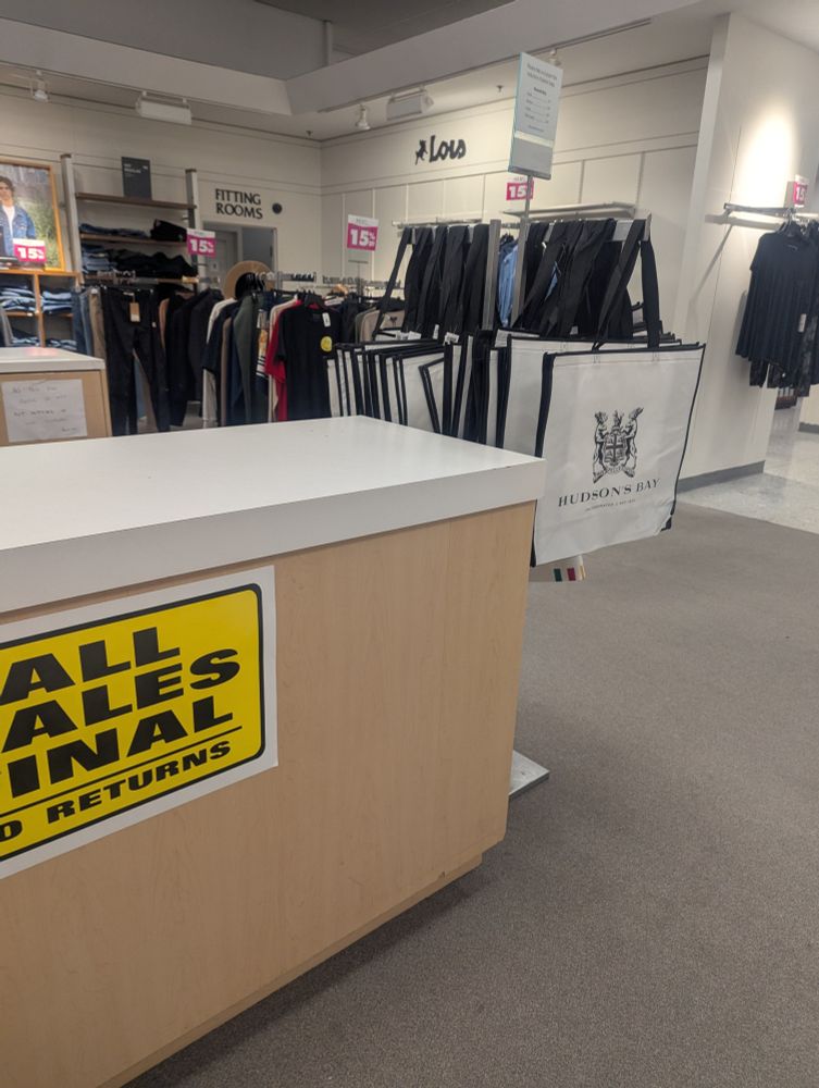 a store - in the foreground a beige counter with white countertop has a yellow sign stuck to it that says "ALL SALES FINAL no returns"
Hudson's Bay branded tote bags hang from a stand to one side 
In the background are racks of clothes and the entry to fitting rooms 