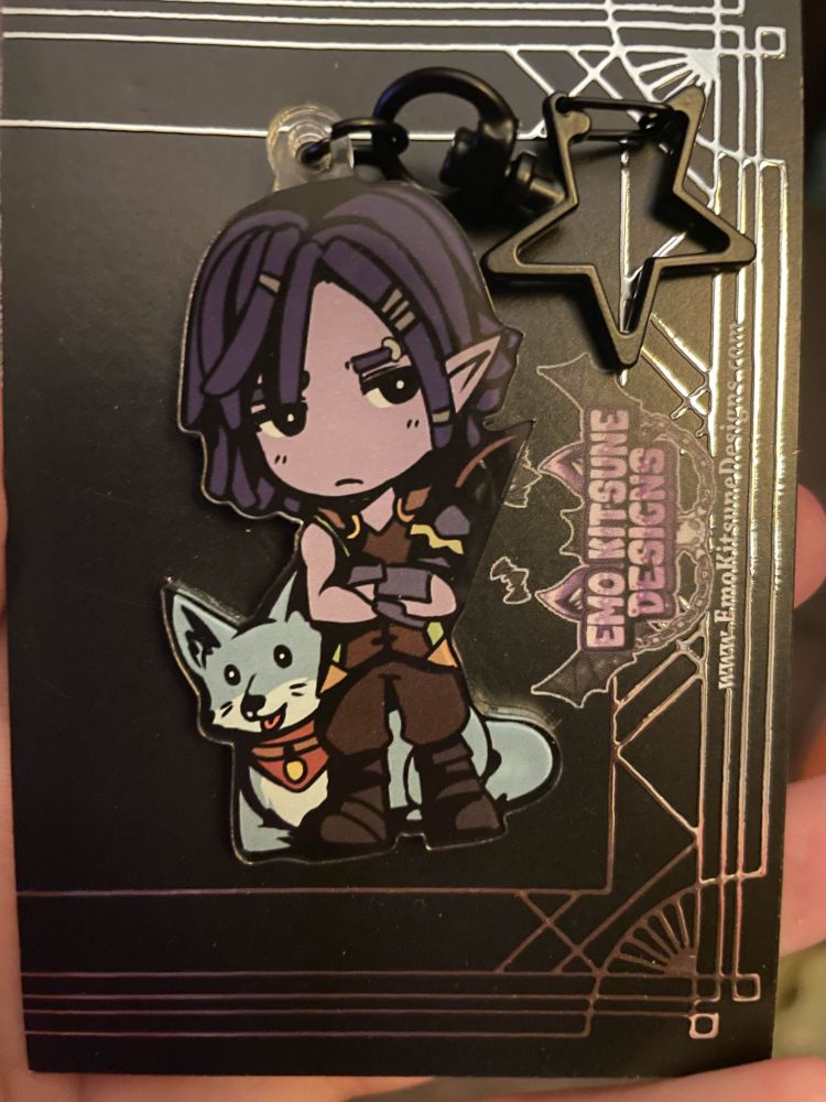 there's an acrylic charm of the character hassian from palia. tau (his pet dog) is behind him. he is attached to a star shaped keyring clip. the back of the charm is attached to a black and silver card backing that says 'emo kitsune designs' on it.