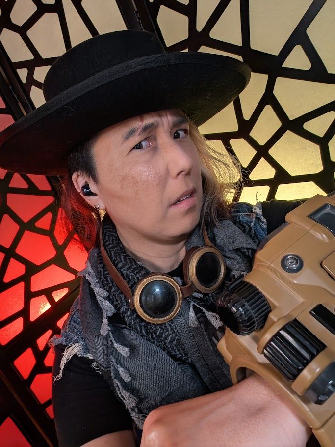 a photo of Cai dressed up for Fallout New Vegas wearing a shreded vest and a Pip-boy (a large, wrist-worn computer from the world of Fallout). He also wears a black, flat-brimmed hat and goggles.