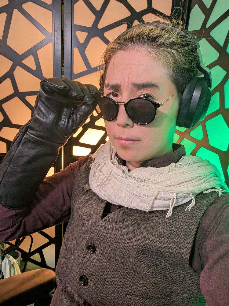 a photo of cai as his character paxton wearing a tweed vest and red shirt with a white scarf. he's wearing sunglasses and leather gloves.