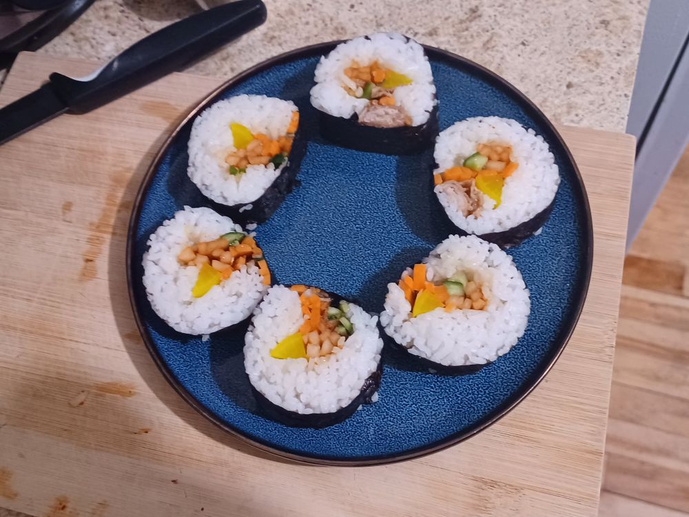 Six gimbap slices on a blue plate. They have orange carrot, green cucumber, yellow pickled radish, pink spicy radish, and brown cooked pork belly inside them.