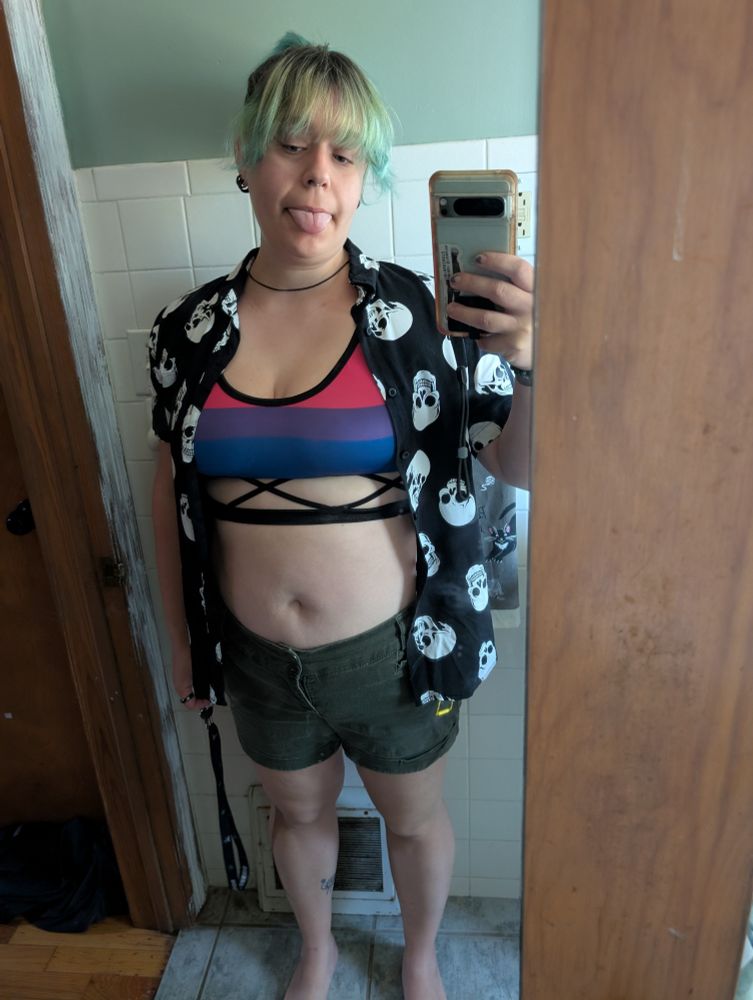A person wearing olive green shorts and a bisexual flag swim top with a skull patterned button down with their tongue out