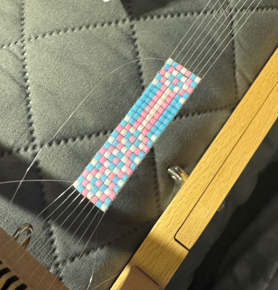 Small seed beads (trans flag colors) in geometric patterns on a bead loom. The bracelet is partially finished