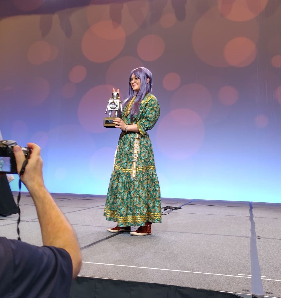 Darcie stands on a stage and holds a little silver robot statue 