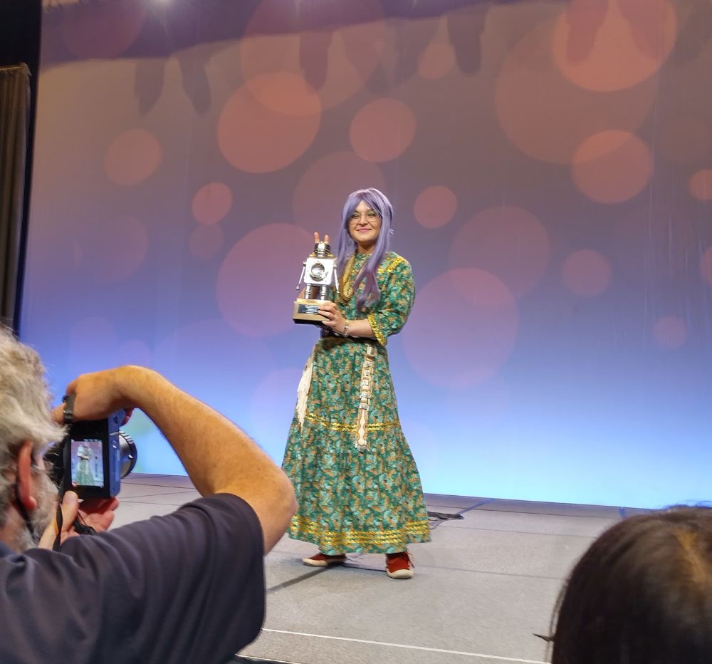 Darcie stands on a stage and holds a little silver robot statue 