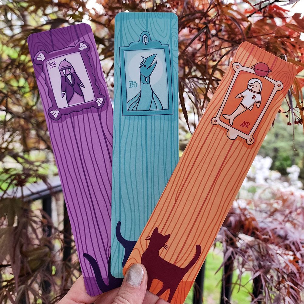 My hand holding three large bookmarks in bright colors against a backdrop of trees.