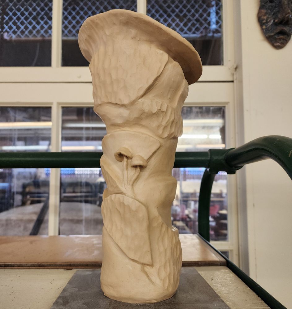 A tall clay vase with mismatched slabs of clay, thin and tall, with some pieces given a nubbly texture and two tiny mushrooms affixed in the middle. The whole things looks like abstract bark.